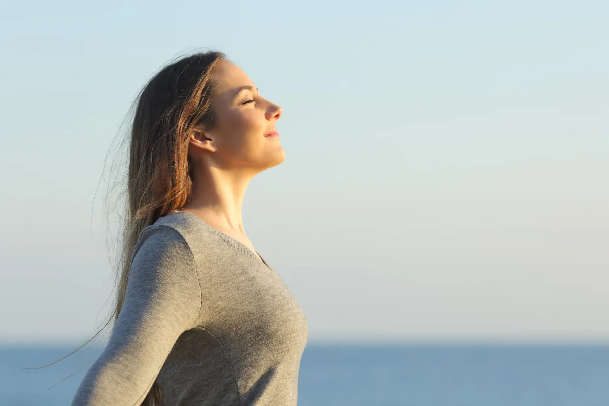 Young woman standing by the ocean with her eyes closed, smiling softly and facing the sun. She appears calm and content, taking a deep breath in a peaceful, coastal setting.