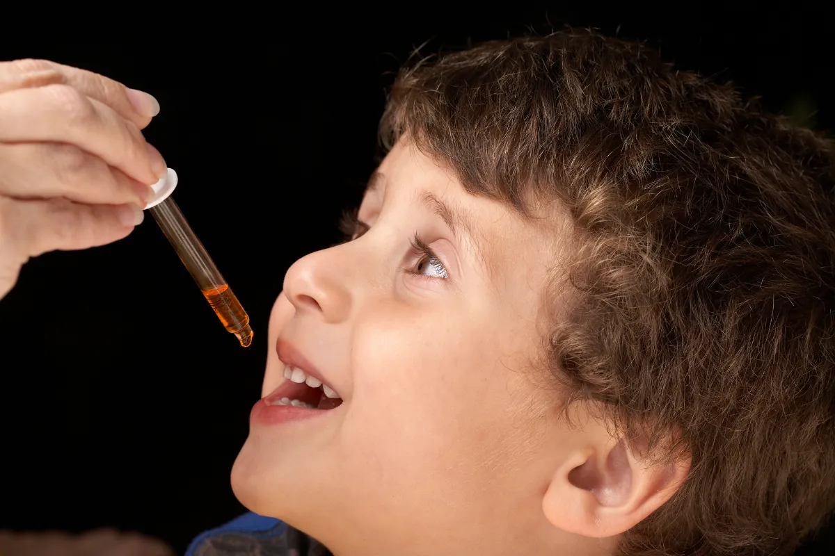 Young child smiling and looking up with mouth open as an adult hand holds a dropper filled with orange liquid, about to administer oral medicine or supplements.