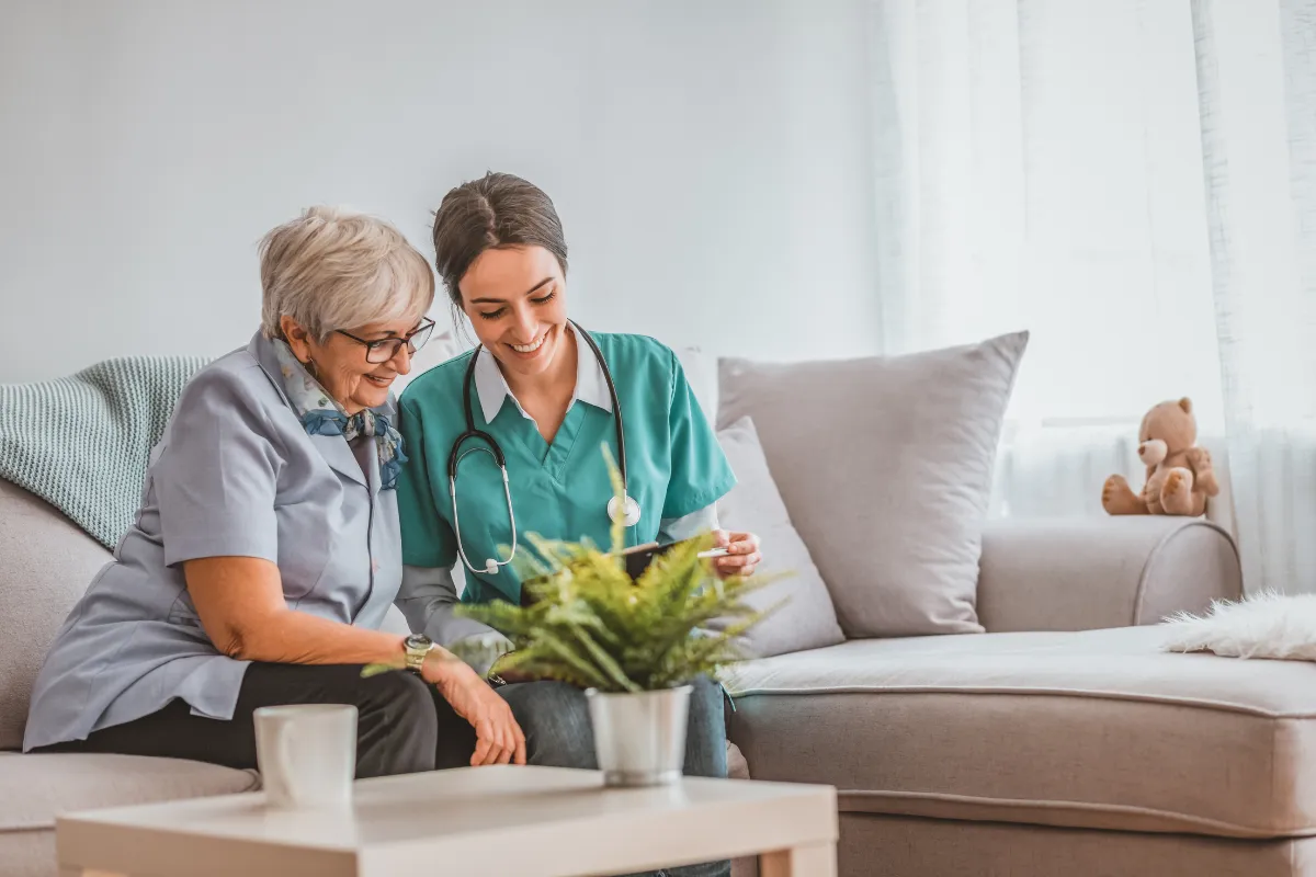Smiling nurse or caregiver in teal scrubs sitting on a couch next to an elderly woman with grey hair and glasses. They are looking at a document or tablet together in a bright, cozy living room, suggesting a home care or health consultation visit.