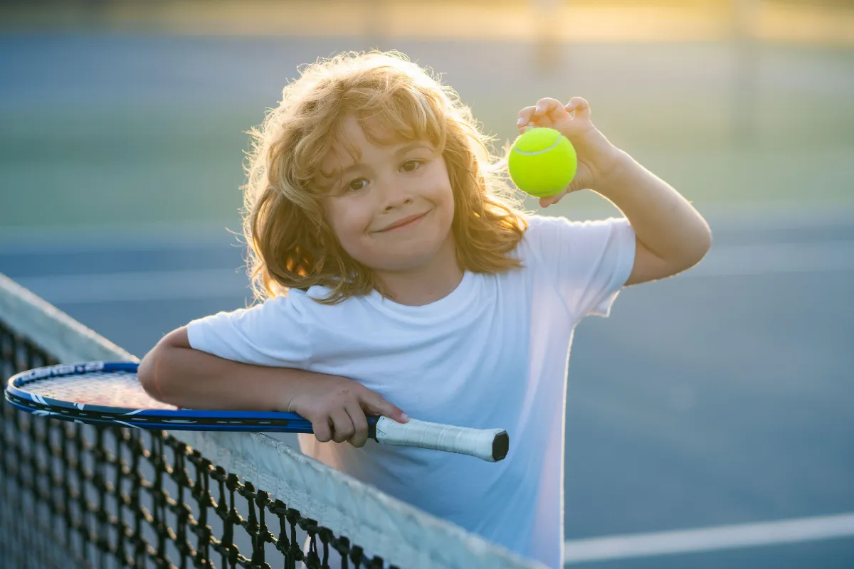 Smiling child with curly blonde hair standing at a tennis court net, holding a tennis ball in one hand and resting a racket on the net with the other. The scene is brightly lit with warm sunlight, suggesting a fun outdoor sports activity.