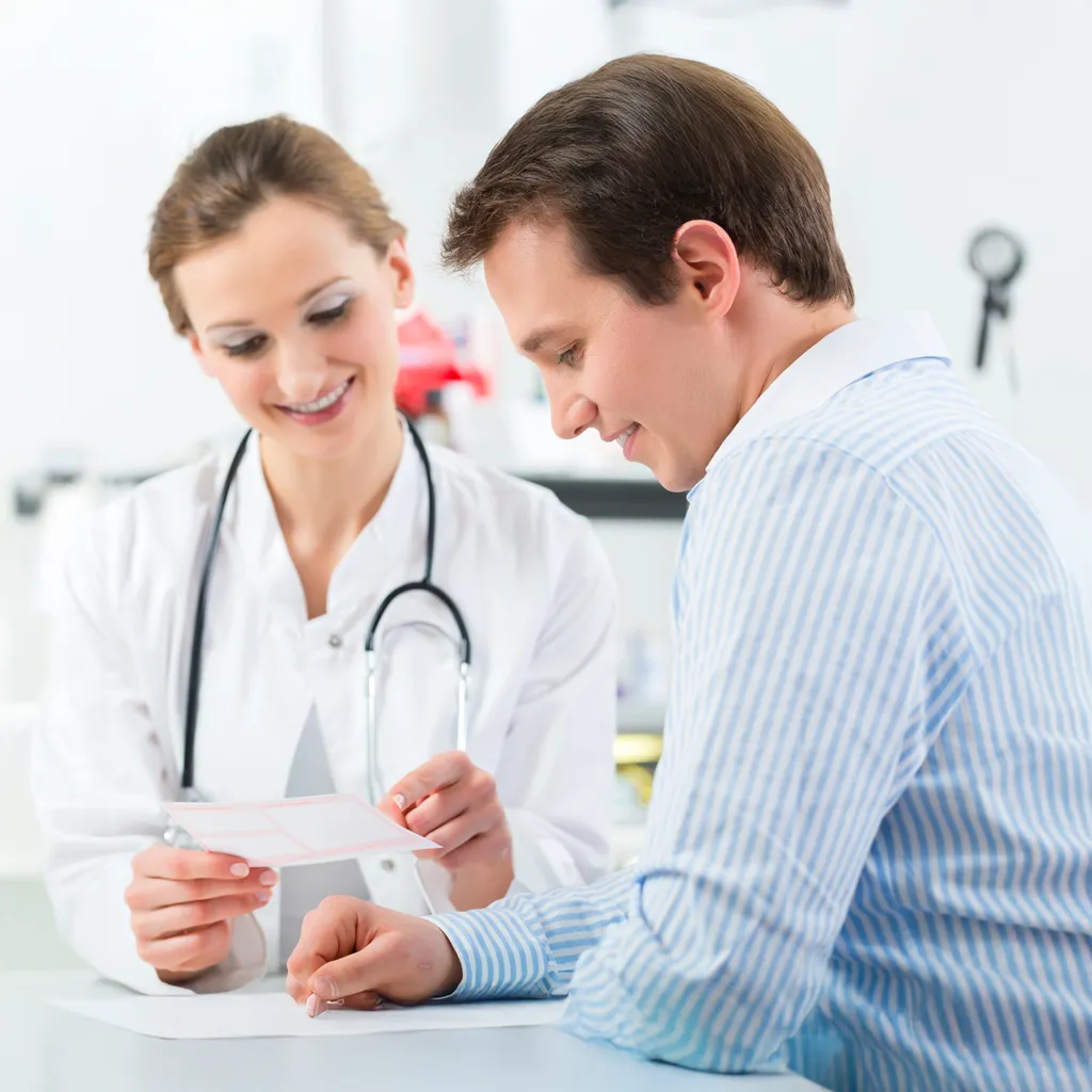 Smiling female doctor showing a medical document or prescription to a male patient in a clinical setting. The doctor is wearing a white coat and stethoscope, and they appear to be having a friendly, informative conversation.