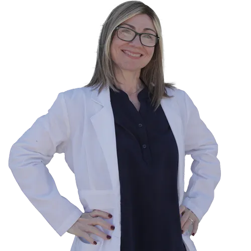 Confident woman with straight blonde hair standing with hands on hips, smiling slightly. She is wearing a dark blouse and a smartwatch, posed in front of a blurred background of a lecture hall with an audience and a speaker on stage.