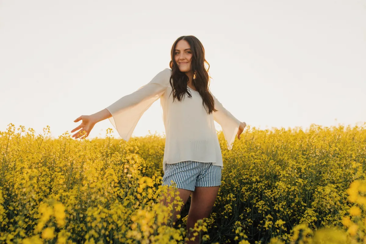 Young woman smiling with arms outstretched while standing in a vibrant yellow flower field under a clear sky. She is wearing a light blouse and striped shorts, enjoying the peaceful outdoor setting.