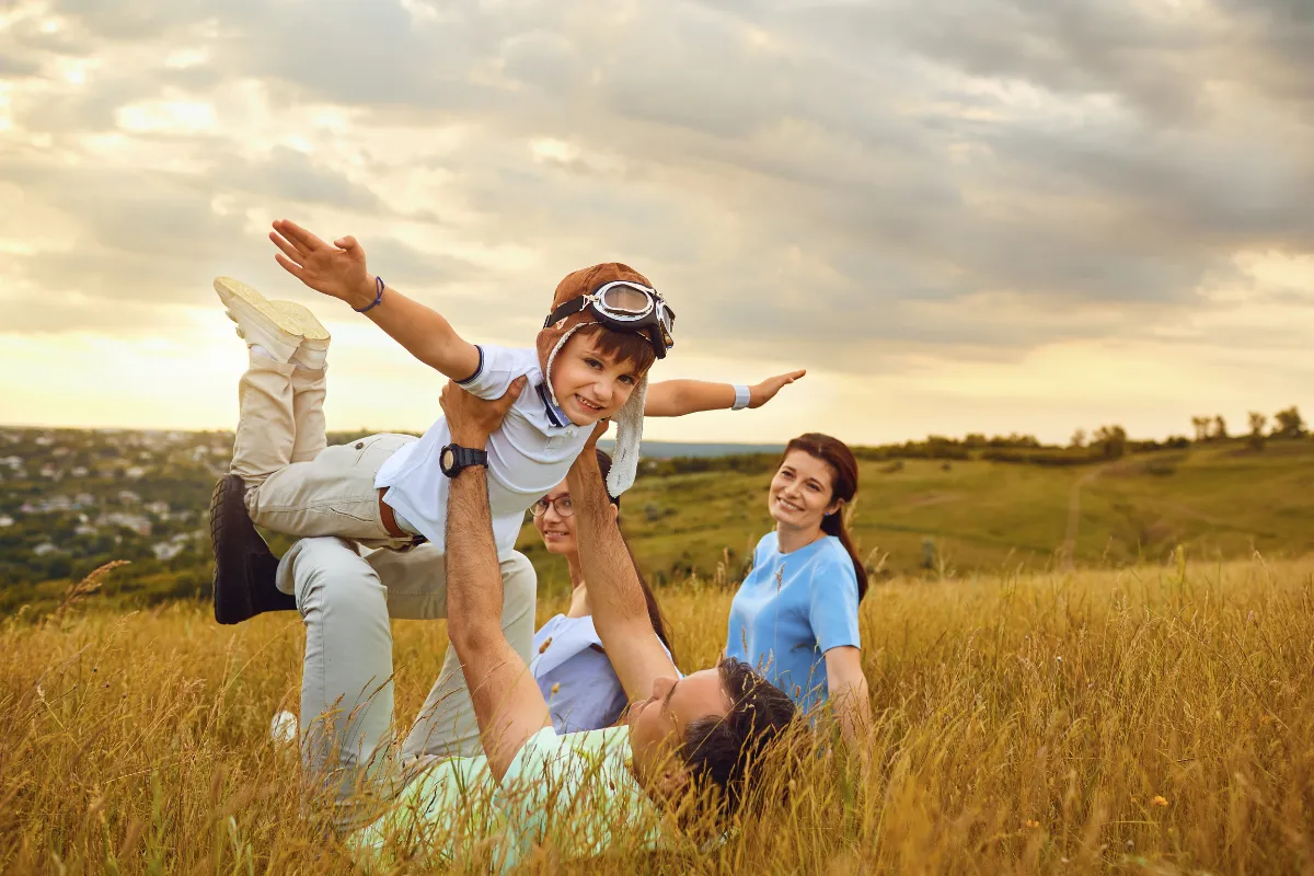 Happy family playing together in a grassy field at sunset. A child wearing aviator goggles is being lifted into the air by an adult, pretending to fly, while two other family members smile and watch. The atmosphere is joyful and carefree.