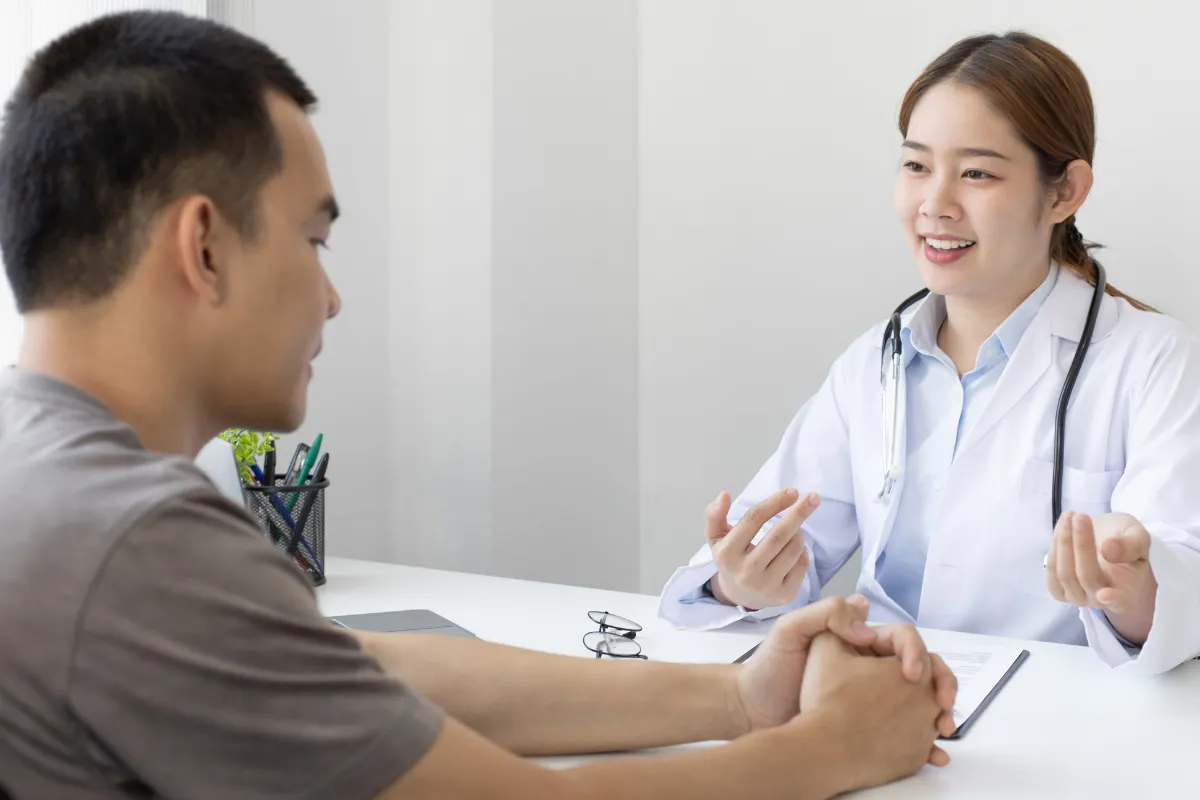 A young female doctor in a white lab coat talks with a male patient in a casual and approachable manner. They are seated across from each other in a well-lit medical office, and both appear engaged in the conversation.
