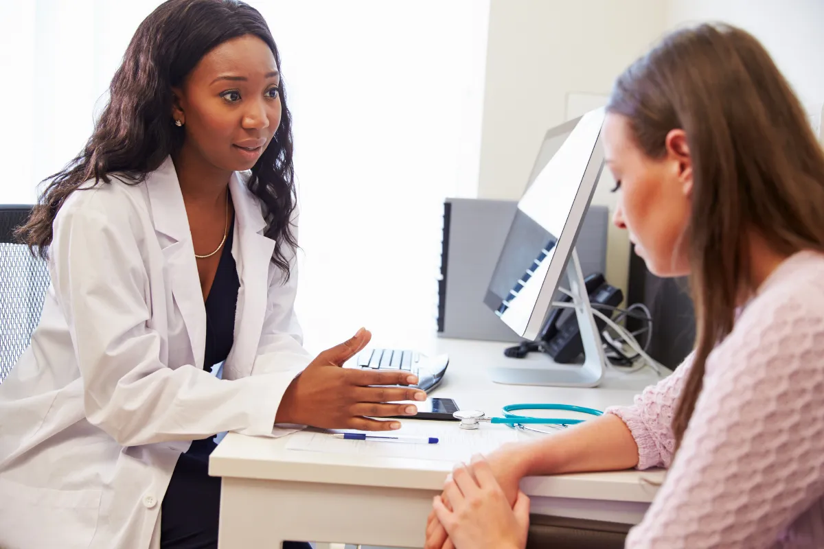 A female doctor with long dark hair, wearing a white lab coat, is sitting at a desk and speaking empathetically to a female patient. The patient is seated across from her, listening attentively. A stethoscope and clipboard are visible on the desk in a bright clinical office.