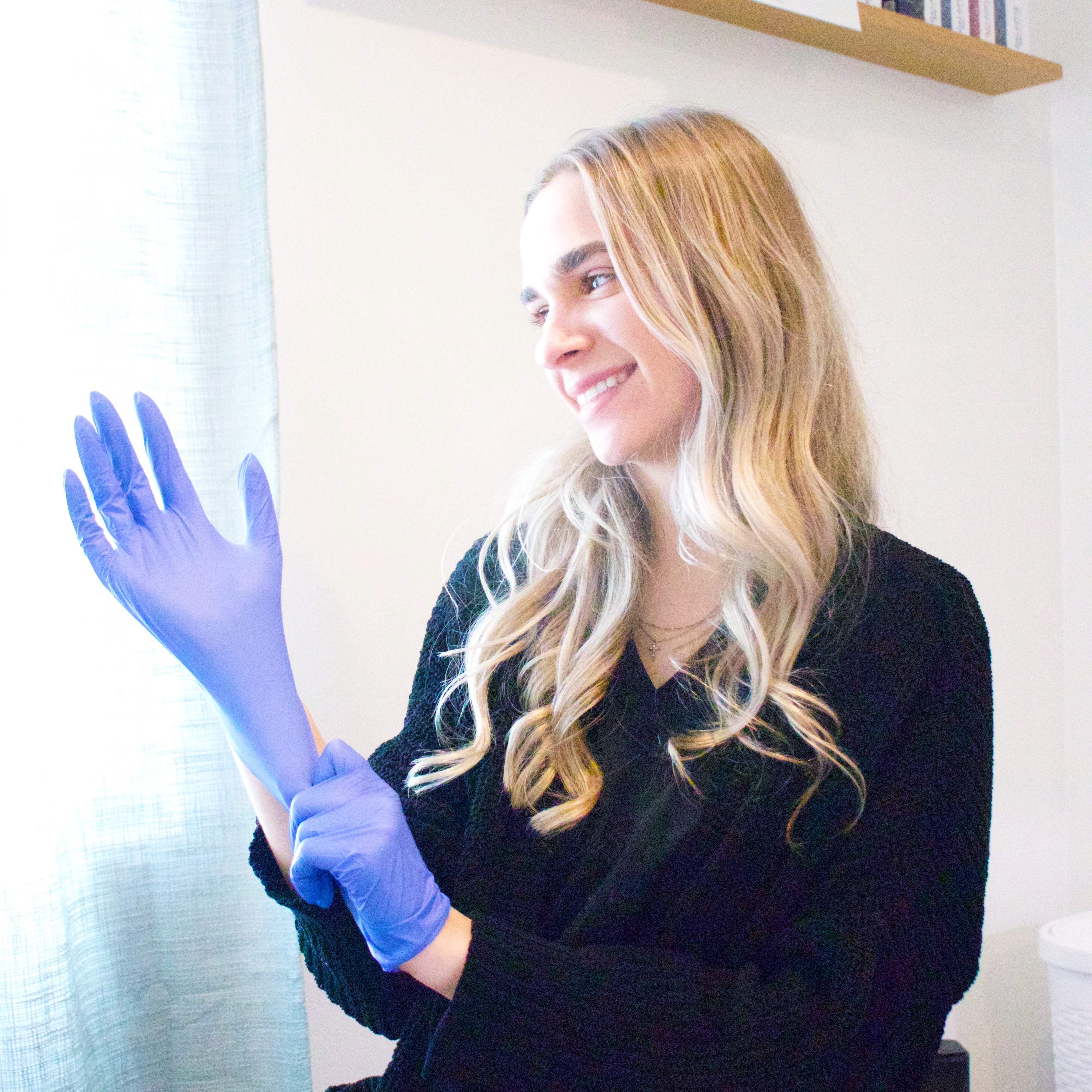 Young child with mouth open, receiving liquid medicine or supplement from a dropper held by an adult. The child is indoors, and the background shows a window with soft daylight and a pink flower pot.