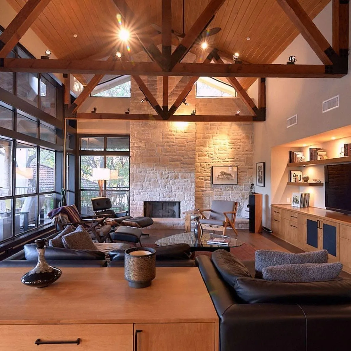 Modern remodeled living room with exposed wood beams, stone fireplace, large windows, and custom built-ins.