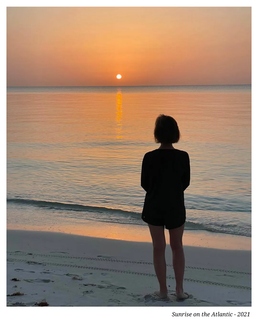A woman standing alone at the water's edge, looking out toward a warm sunset over a calm sea.