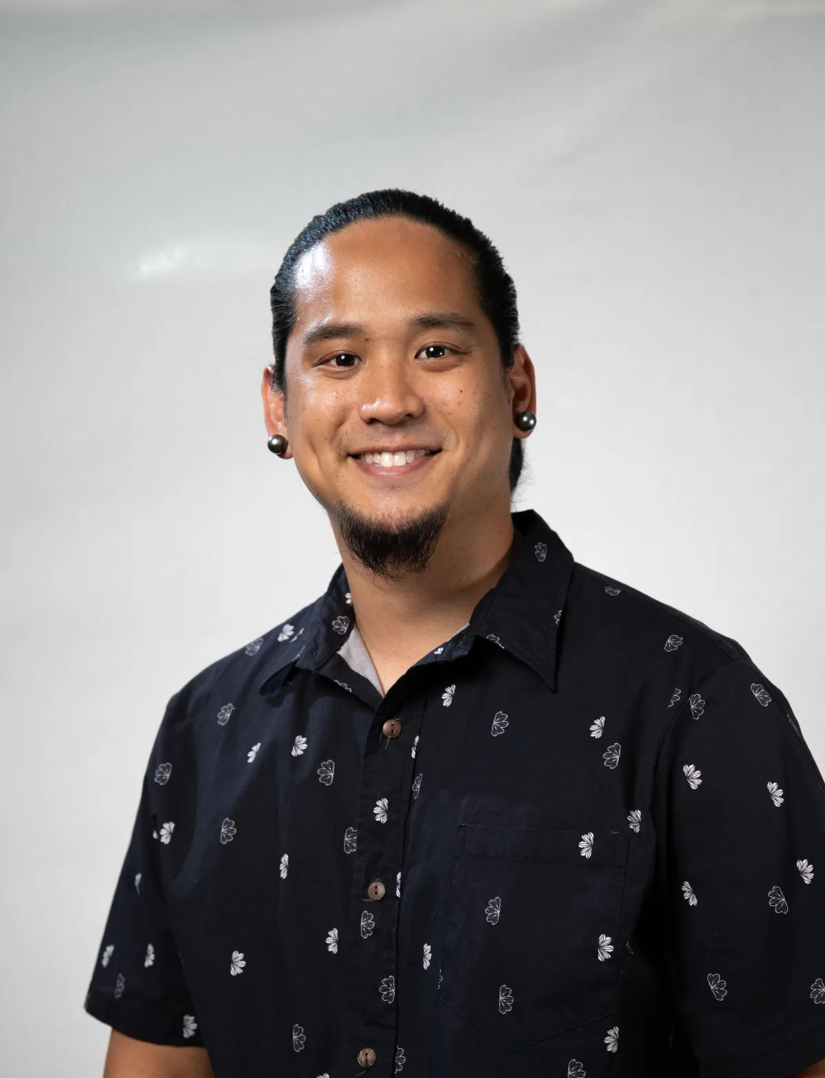 Portrait of a smiling Hawaiian Lomilomi therapist in traditional attire, standing in a softly lit studio with woven mats and tropical flowers. The therapist exudes warmth and expertise, inviting trust and comfort in a peaceful, welcoming environment.