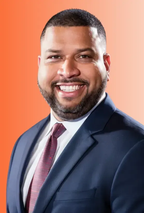 A portrait photo of a Latino man with a beard and mustache, smiling and looking directly at the camera. He is wearing a dark blue suit jacket, a white collared shirt, and a patterned red tie. The background is a solid, warm orange color.