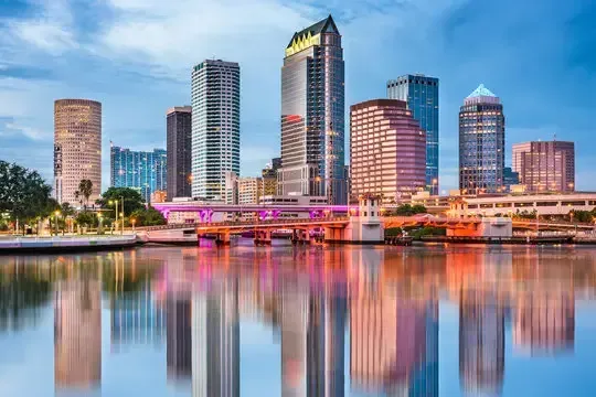 A wide-angle, eye-level shot captures the stunning downtown Tampa skyline reflecting beautifully on the calm water of the Hillsborough River at dusk. The city lights are just starting to glow, creating a beautiful contrast against the soft, pastel-colored sky of blue, pink, and purple. The modern high-rise buildings, including the prominent cylindrical Rivergate Tower and the rectangular skyscrapers with their distinctive glowing windows, stand tall and are perfectly reflected in the water below. A couple of bridges, including the Cass Street bridge, are visible crossing the river in the foreground. This image is a great example of local SEO Tampa content, showcasing a classic and picturesque view of the city from the water. It highlights the vibrant and modern feel of downtown Tampa.