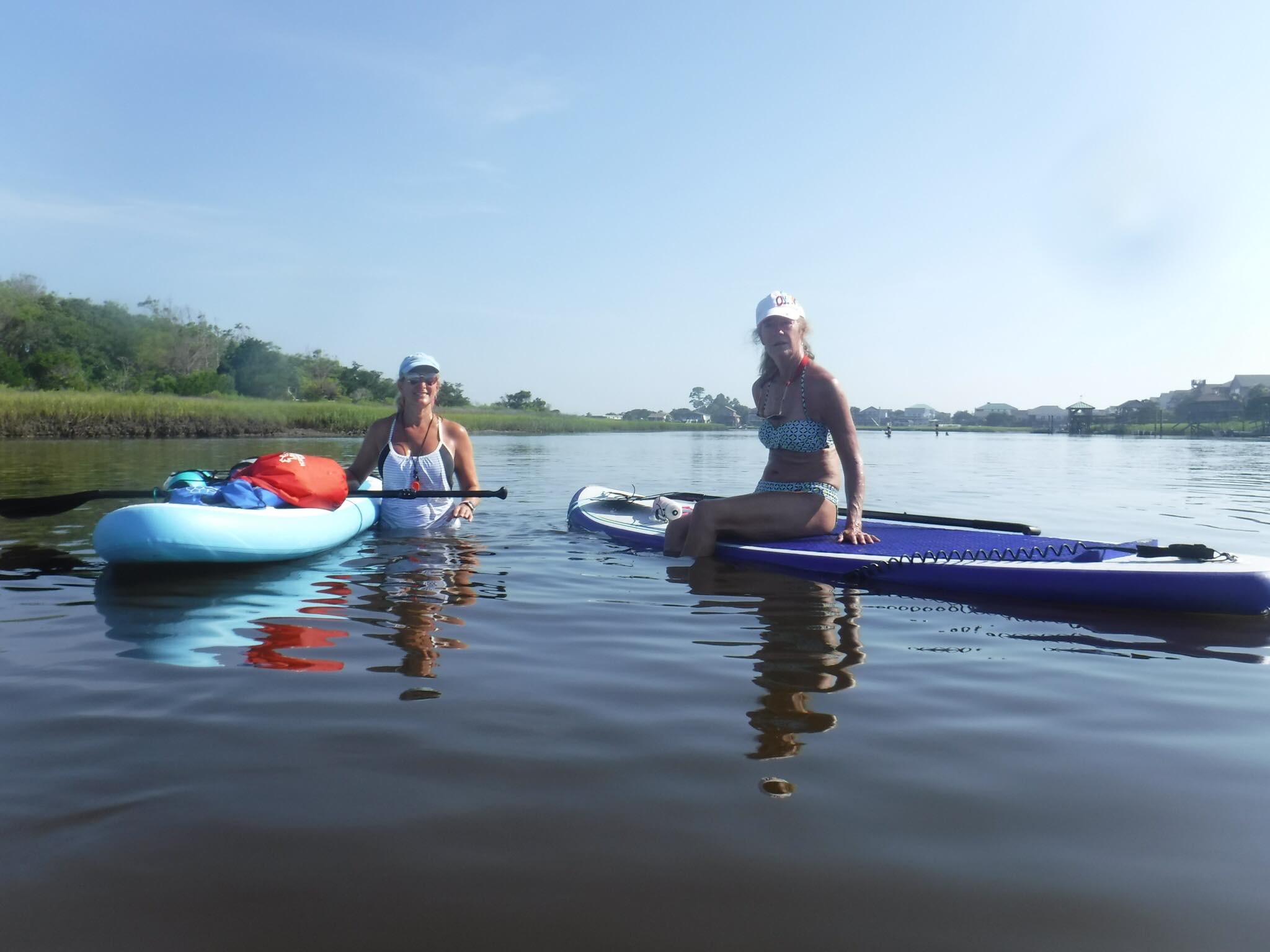 two women one sitting on a paddleboard in the water and one standing in the water holding on to a paddleboard