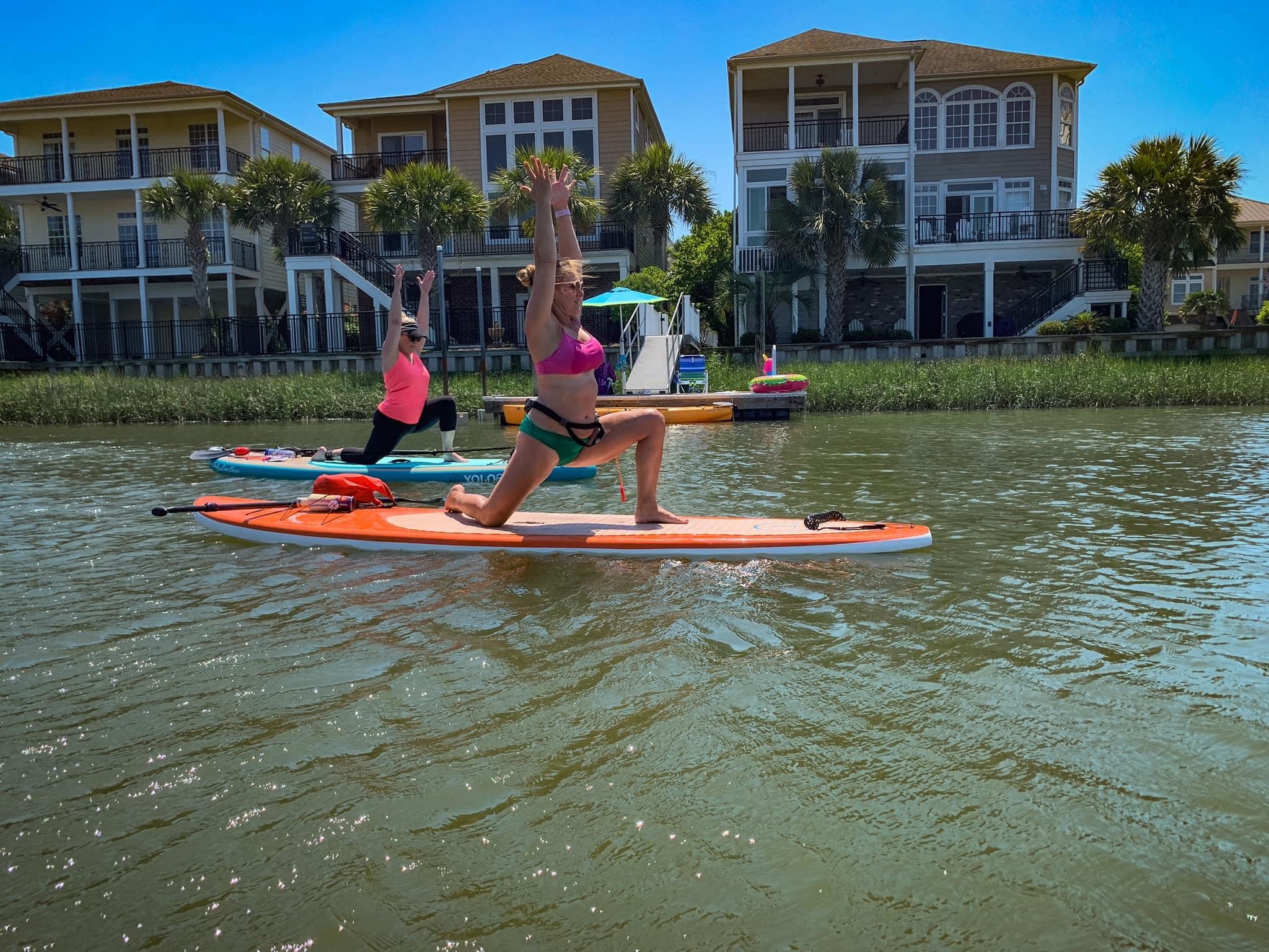wome doing yoga poses on paddleboards on the water with homes in the background