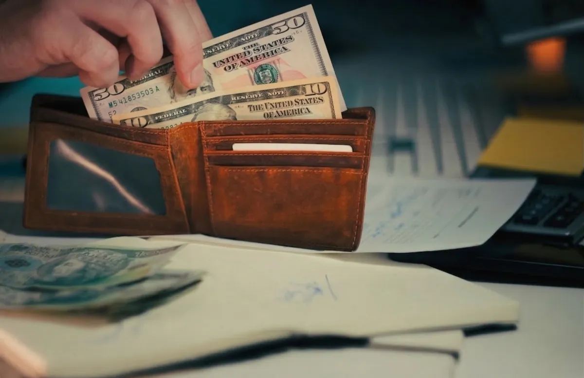 Hand placing dollar banknotes into a brown leather wallet.