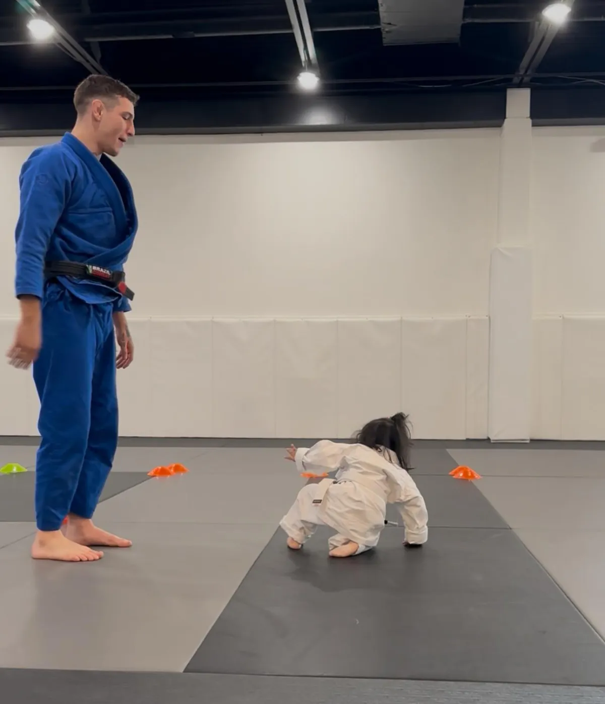 Child in white BJJ gi practicing movement on mat, instructor in blue gi guiding, colorful cones in background, emphasizing fun and fundamentals in kids' Brazilian Jiu-Jitsu class for ages 3-5.