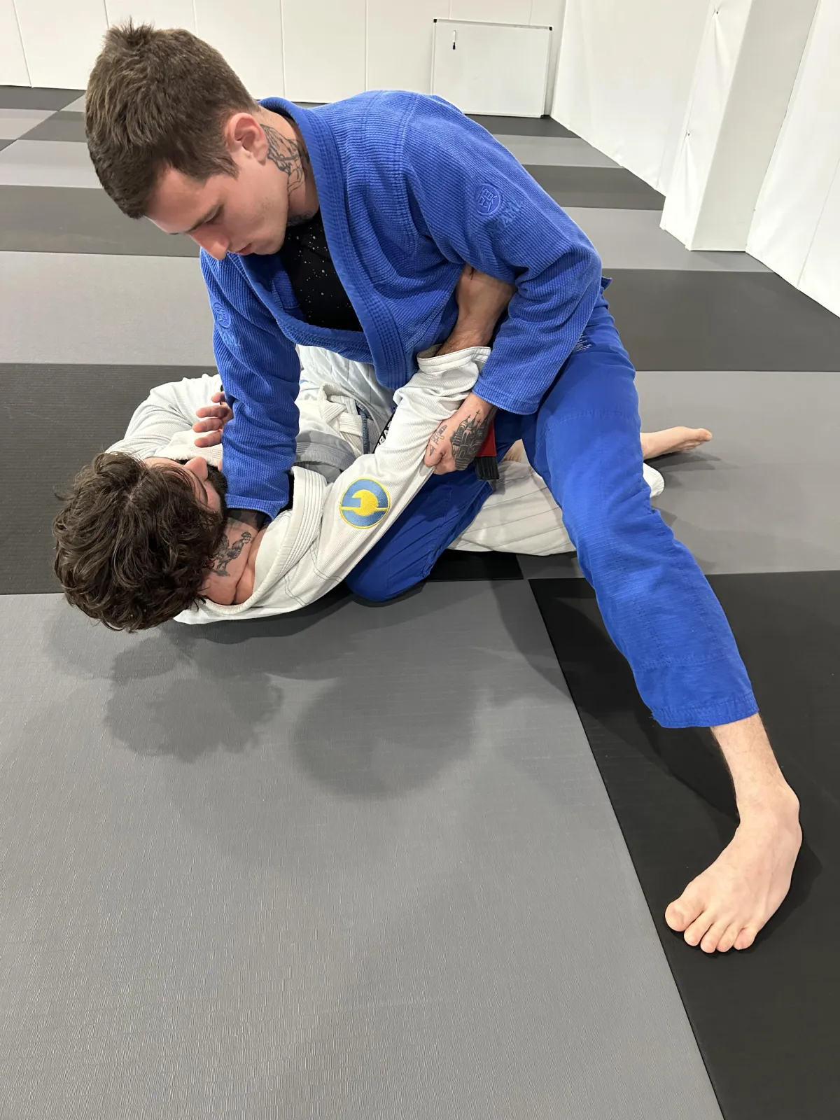 Two martial artists practicing Brazilian Jiu-Jitsu on training mats, one in a blue gi applying a technique on the other in a white gi, demonstrating skill development and engagement in a martial arts environment.