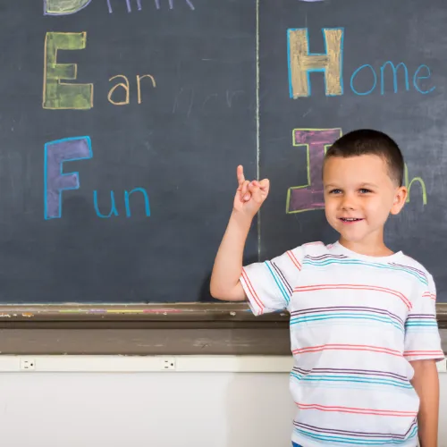 A young boy with a big smile stands in front of a chalkboard displaying colorful letters and words, promoting a supportive learning environment for diverse student needs.