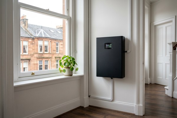 A SolaX Power X1 hybrid inverter (white unit) and a SolaX Triple Power 5.8kWh battery storage unit (dark grey/black unit) mounted side by side on a white interior wall. The battery display shows 92% charge and the status "ACTIVE" in green. Black cables connect both units down to a grey consumer unit with red circuit breakers labelled Main, Battery, PV, and House. A wooden sash window with a small succulent plant on the sill lets in natural daylight. Traditional red sandstone buildings are visible through the window.