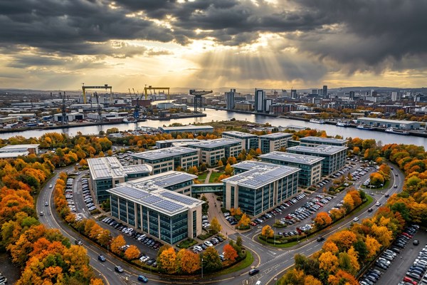 A dramatic aerial drone photograph of a large modern commercial business park, likely in Glasgow, Scotland. Several multi-storey glass office buildings with flat rooftops are covered in large arrays of solar photovoltaic panels. The complex is surrounded by full car parks, autumn-coloured tree-lined pathways, and green landscaped areas. Industrial warehouses, railway lines, shipping containers, and port infrastructure are visible in the mid-ground, with the River Clyde and city skyline in the distance. Dramatic sunbeams break through dark storm clouds overhead.
