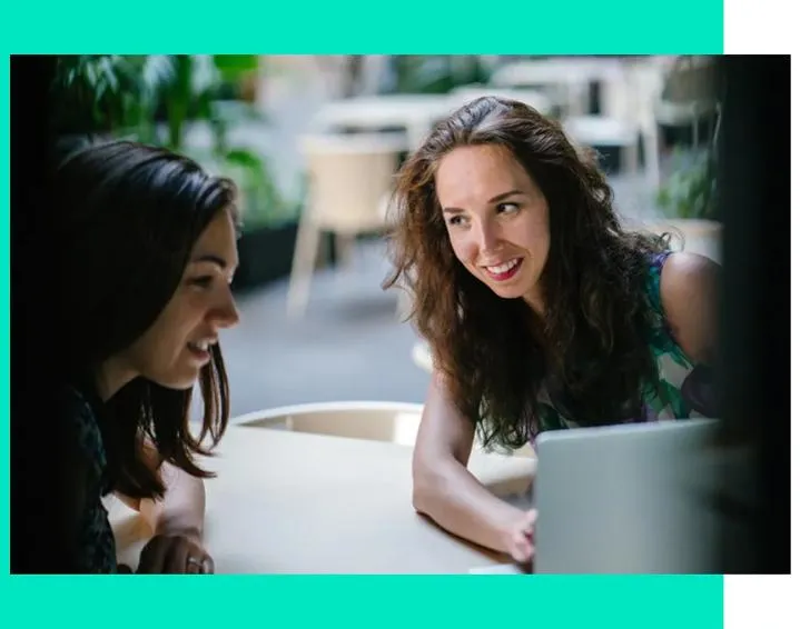 Two women at a table with a laptop, immersed in discussion, surrounded by a refreshing green background