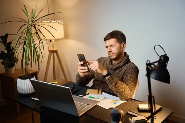 A man at a cluttered home desk pauses his work to check his phone, surrounded by charts, a laptop, and warm lamp light.