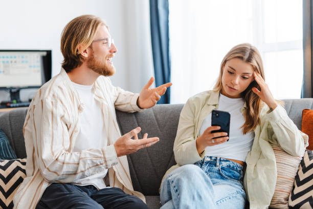 A man talks animatedly on a sofa while his partner looks away at her phone, appearing distracted and overwhelmed.