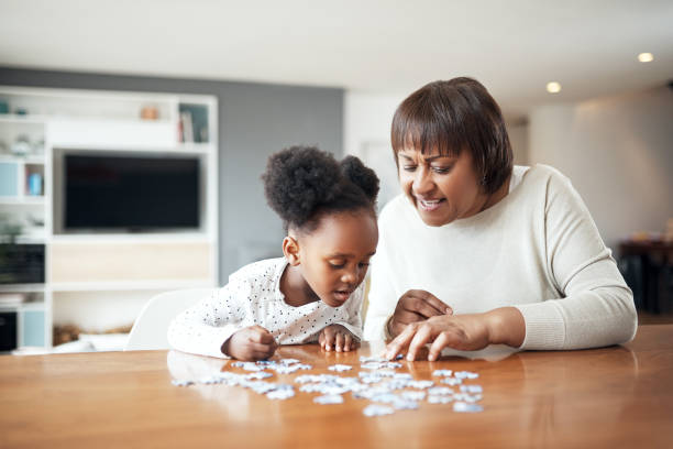 A young girl leans forward intently over puzzle pieces on a table while a woman beside her smiles and helps guide the activity in a bright, relaxed living room.