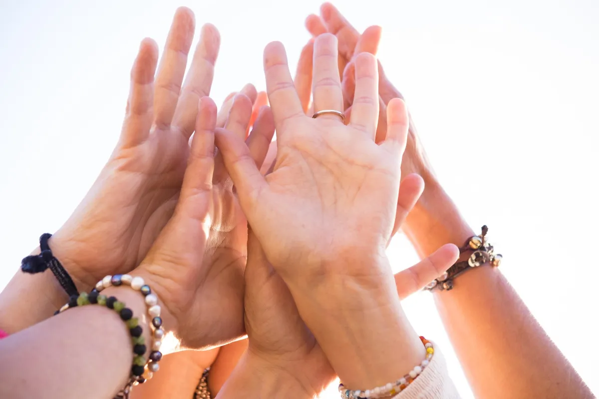 Hands of diverse individuals reaching upward, symbolizing hope and unity against a bright sky backdrop.