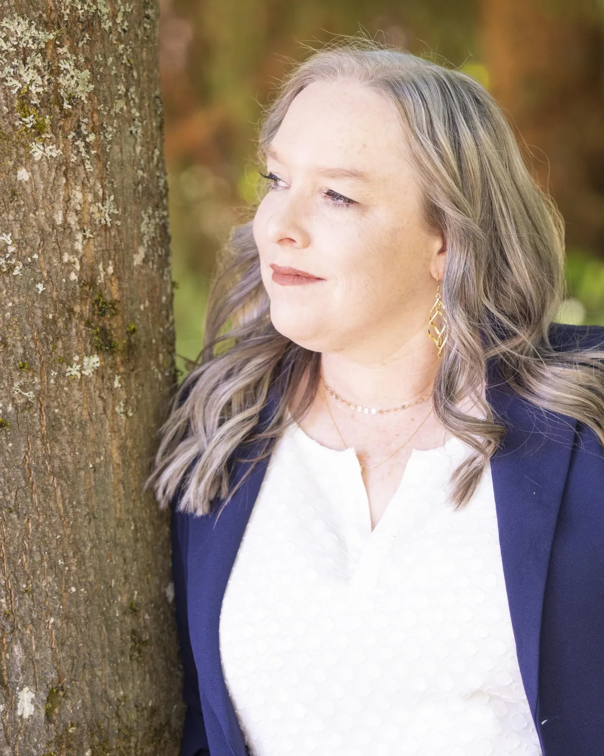 Karen Nelson leans against a tree outdoors, looking to the side with a calm expression. She wears a navy blazer, white textured blouse, and gold jewelry, with sunlight filtering through the background.