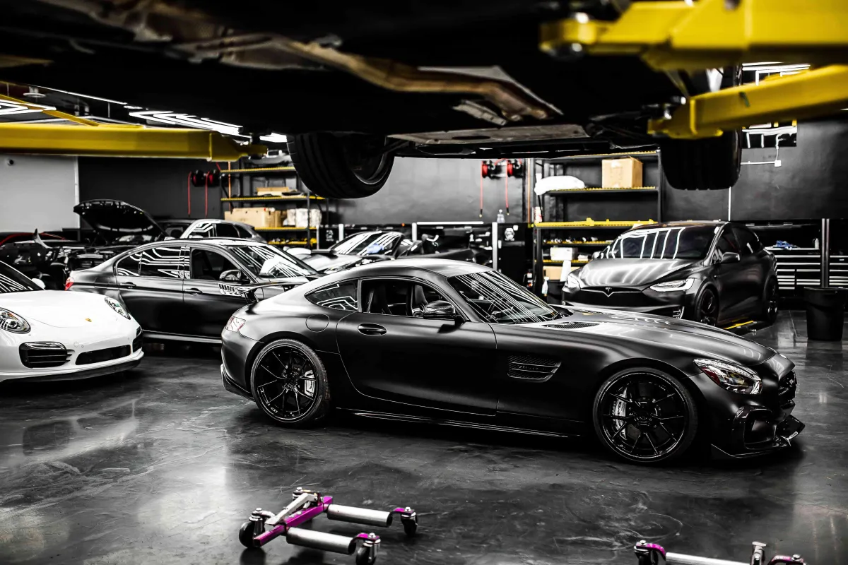 a while porsche, black mercedes gt, black tesla model x, with many other cars parked inside a auto service repair shop 