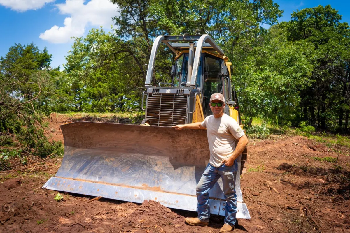 Experienced land clearing contractor in Oklahoma standing next to a John Deere dozer on a cleared job site surrounded by trees