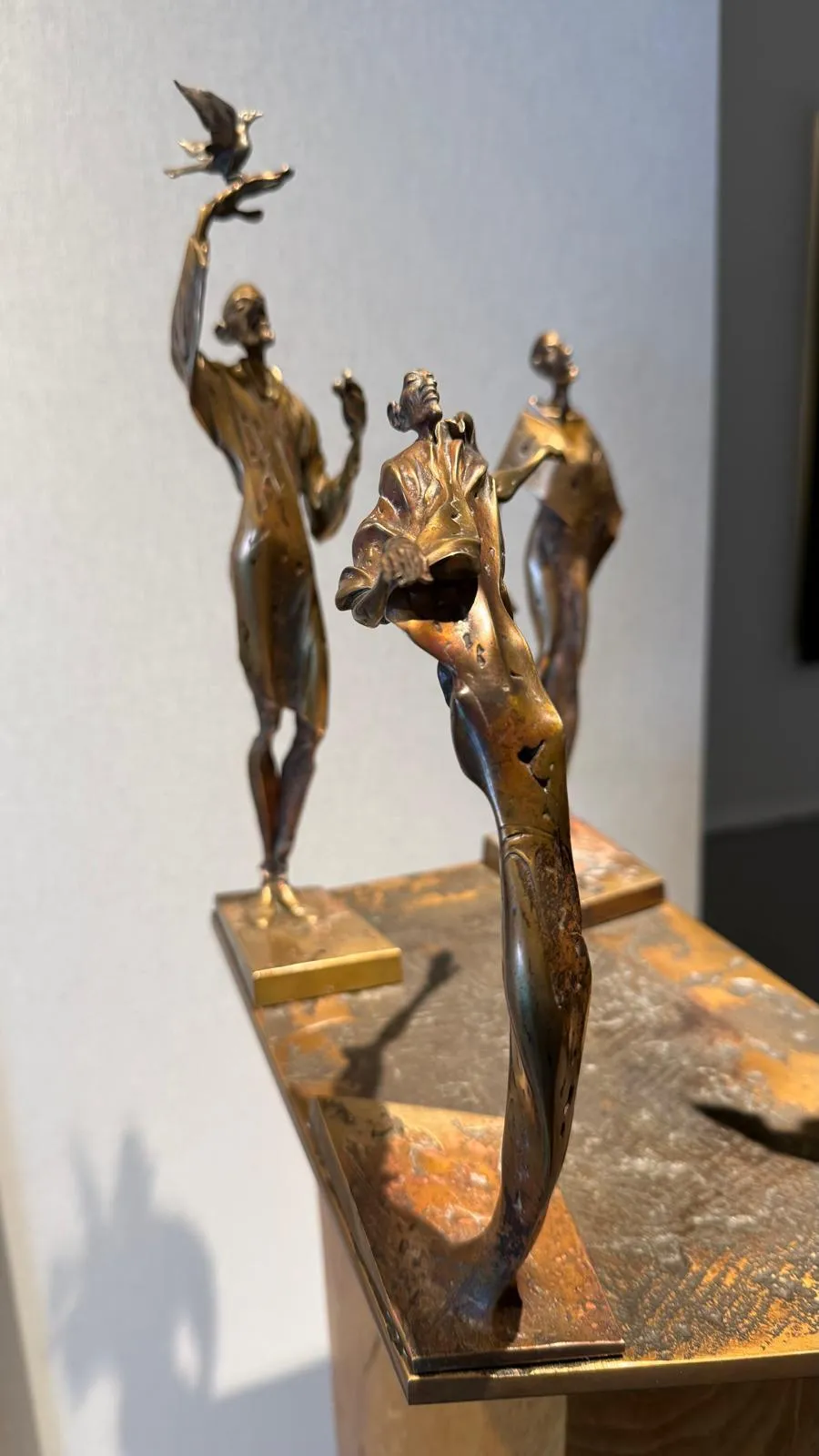 Vordere CuSi3 Siliziumbronze-Skulptur „Herbst“ (ca. 25 cm): schlanke Figur in starker Rückbiegung mit anliegendem Gewanddetail, patinierte Oberfläche in warmen Bronze- und Brauntönen, auf Metallplatte vor hellgrauer Galeriewand; zwei weitere Figuren unscharf im Hintergrund.