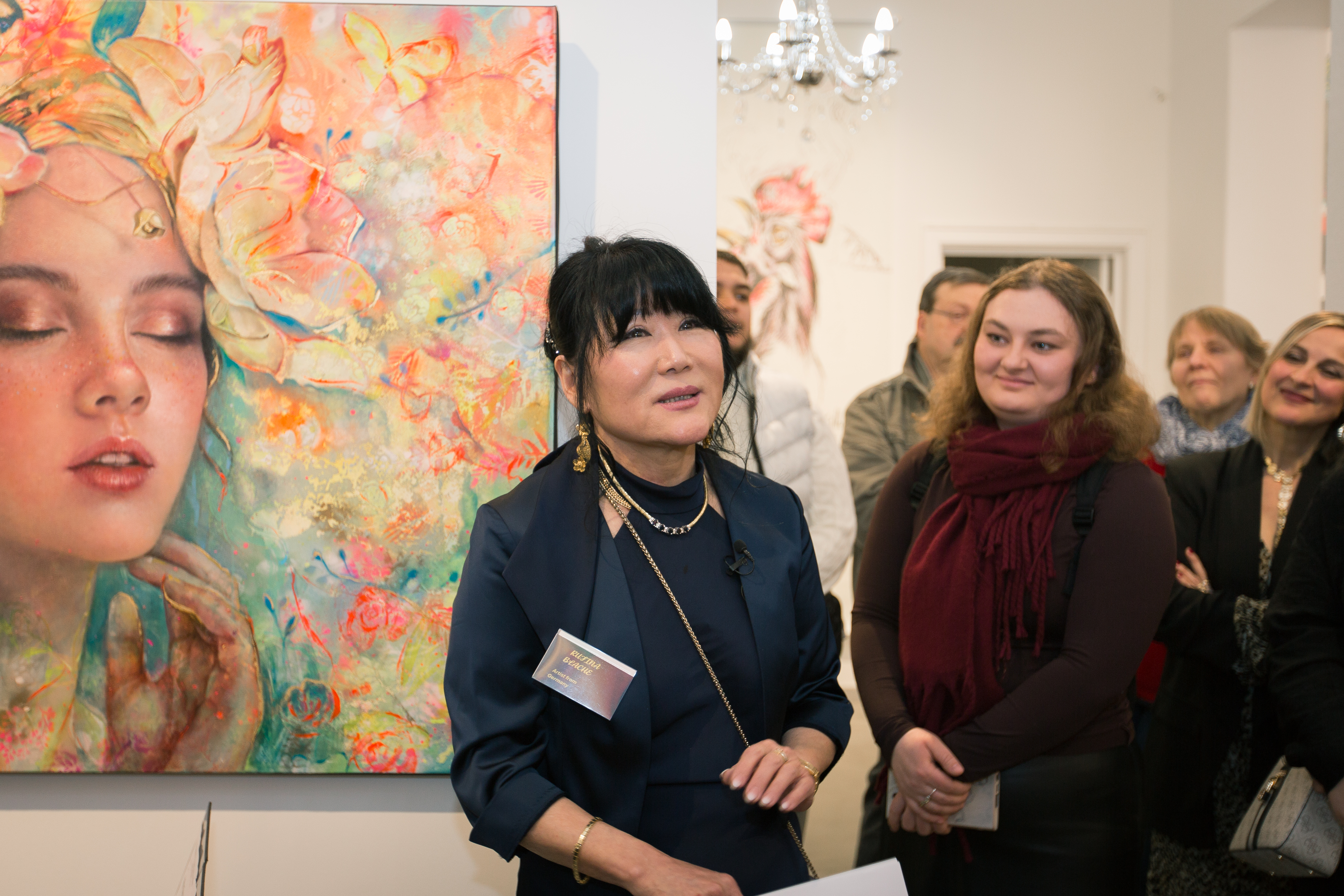 A close-up of a contemporary artist's hands delicately applying gold leaf to a large, textured canvas. The studio background is softly blurred, with warm, natural light and subtle copper reflections. The scene conveys craftsmanship, focus, and the tactile process of creation.