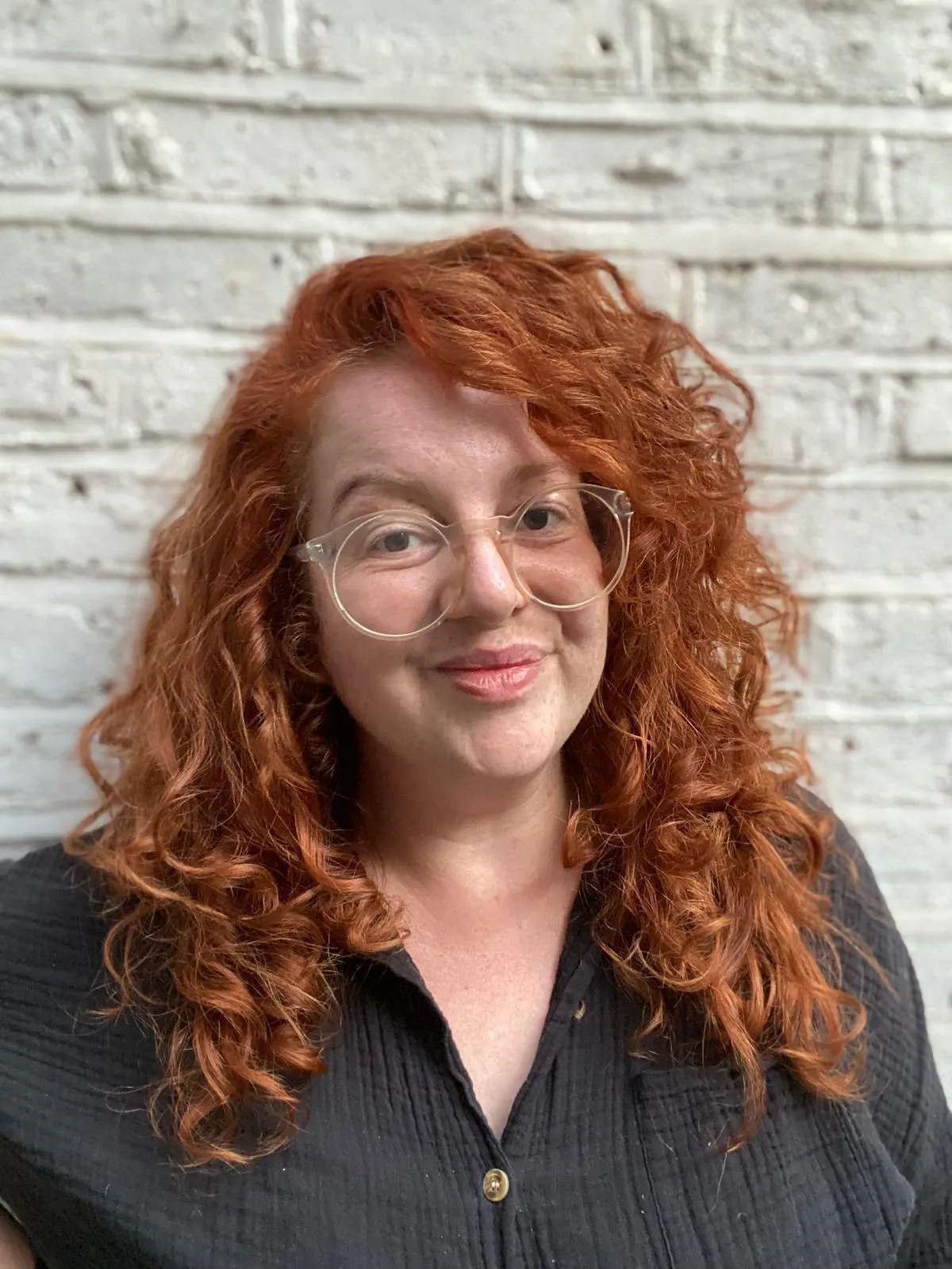 A white woman with long curly red hair, large round transparent glasses and a black shirt, smiles closed-mouthed to camera. She stands in front of a painted white brick wall. 