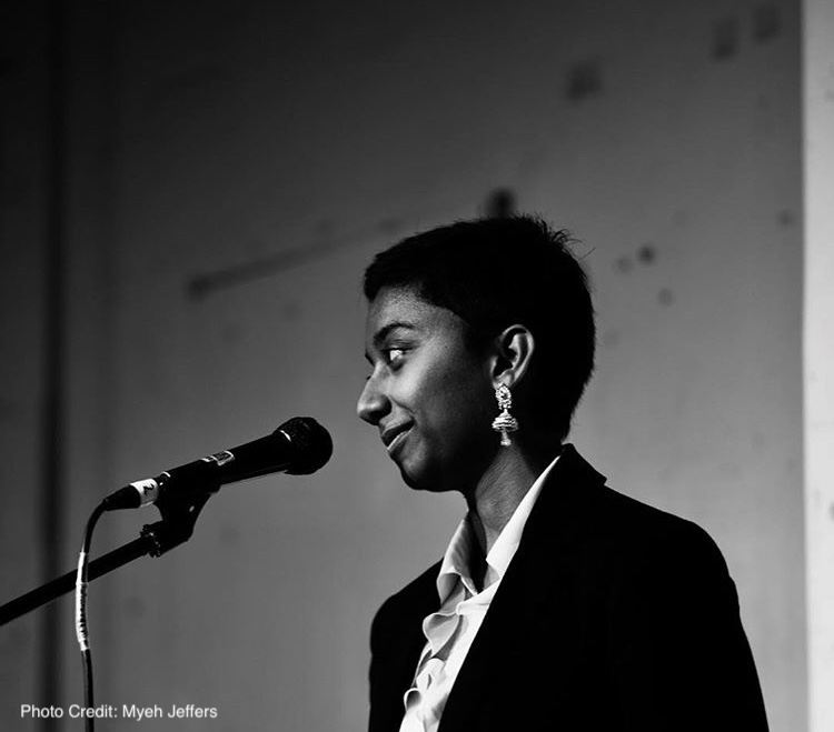 A monochrome photograph of Divya, a woman of South-Asian descent with short dark hair, mushroom-shaped earrings, and who wears a black blazer over a white shirt. She is speaking into a microphone, while smiling slightly, and looking away from the camera. 