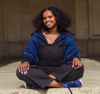 A photograph of Warda, a Black woman of East African descent, with shoulder-length curly hair. She wears a loose blue hooded jumper under a pair of black dungarees, paired with blue trainers. She smiles open-mouthed direct to camera as she sits cross-legged in what looks like an old stone building. 