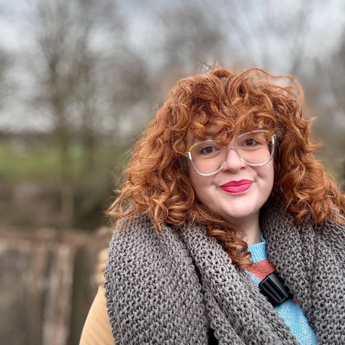 A photo of Zara, a white woman with curly red hair and oversized transparent boxy glasses. She smiles closed-mouthed into camera, while wearing a large grey scarf over a beige coat and multi-coloured jumper. 