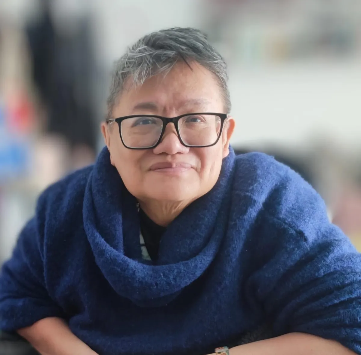 A colour image of Eleanor, a woman of South East Asian descent, with short grey hair and dark plastic-rimmed glasses.. She wears a dark blue cowl neck jumper, and smiles closed-mouthed straight into the camera. 