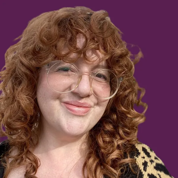 Zara, a white woman with curly red hair and oversized glasses, smiles closed-mouthed to camera. c