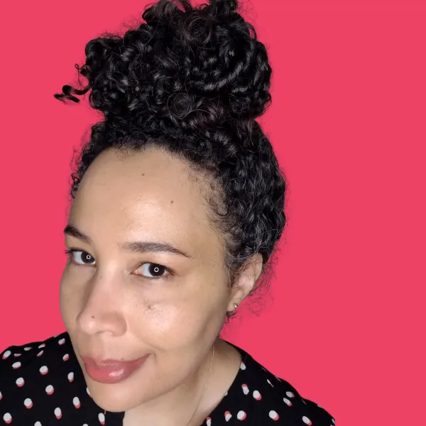 Helene, a Black woman with long curly hair in a bun on top of her head, and a polka dot top, smiles slightly at the camera. 