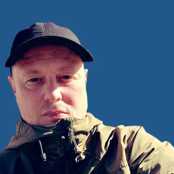 Nick, a white man wearing a navy baseball cap and a parka coat done up to his chin, looks at the camera.