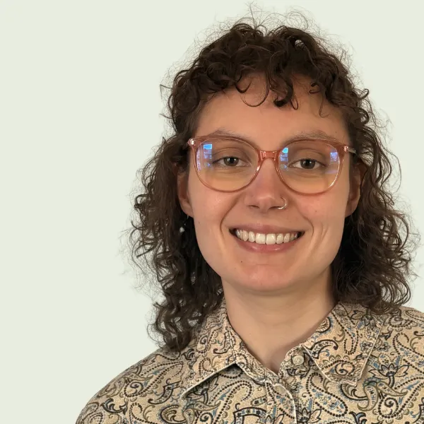 Lauren, a white woman with curly brown hair, large glasses and a light-coloured paisley shirt, smiles to camera. 