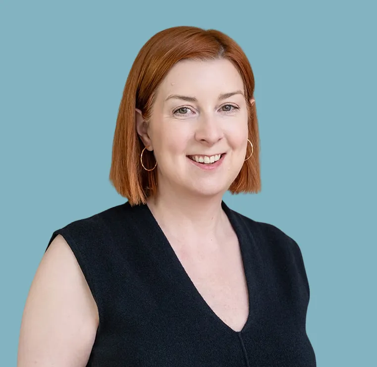 A headshot of Karen, a white woman with straight red hair in a precise chin-length bob, wearing small hoop earrings and a black top, smiling to camera. The background is a kind of duck egg blue.