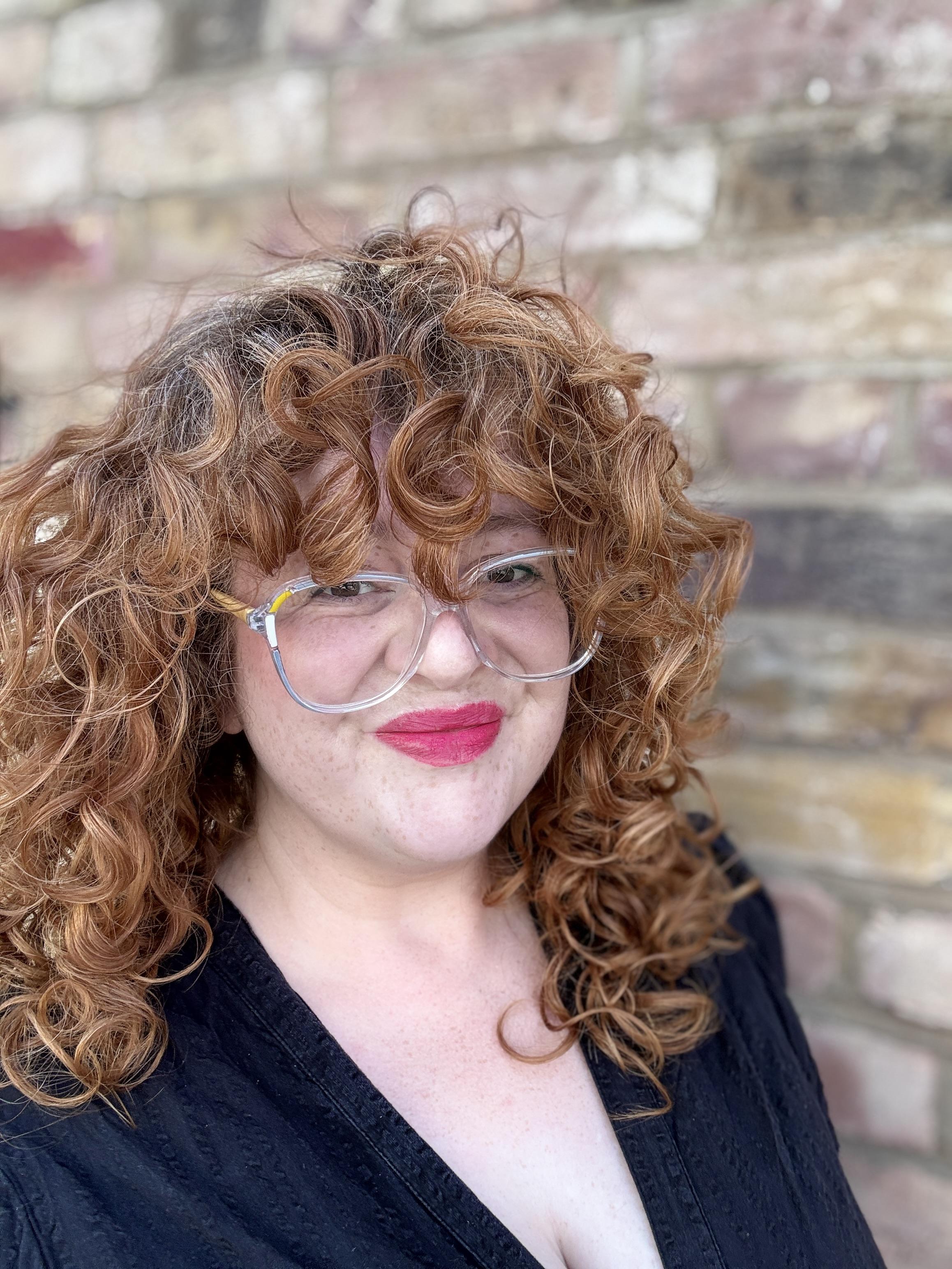 Dr Zara Bain, a white woman with curly red hair, oversized translucent plastic-framed glasses with yellow and white accents, and pink-red lipstick, smiles to the camera. She is wearing a black dress with a deep V neck. Behind her is a yellow and brown brick wall.