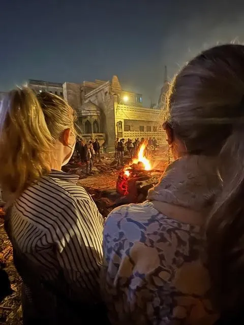 Nighttime scene of an Aarti ceremony with priests holding tall, multi-tiered flaming oil lamps on a riverbank