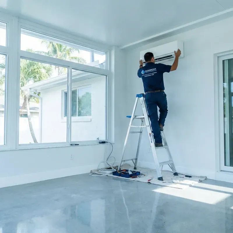 An HVAC technician in a navy blue uniform stands on a silver stepladder inside a bright, modern sunroom. He is mounting a white ductless mini-split indoor unit high on a plain white wall. The room features large floor-to-ceiling windows showing tropical palm trees outside and a polished concrete floor with a protective cloth and toolkit spread out below the ladder.