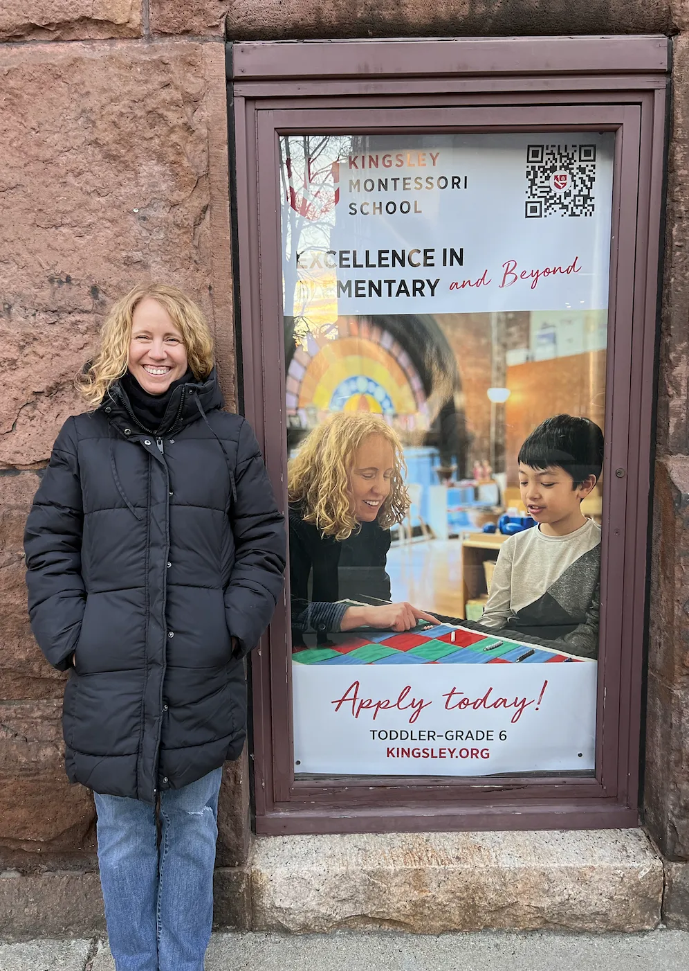 Anne MacMillan standing outside Kingsley Montessori School in Boston in 2023 beside a school window advertisement showing her teaching a Kingsley elementary student multiplication with the Montessori checkerboard, an image used by the school in its 2023 promotional materials.
