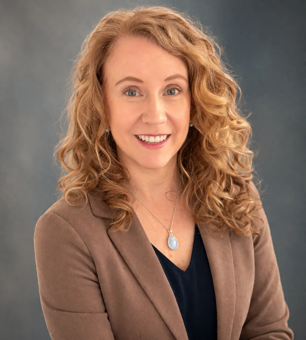 Professional headshot of Anne MacMillan wearing a brown blazer and blue pendant necklace she uses to represent her autistic little brother against a neutral studio background.