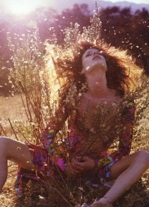 Bohemian woman in a vibrant floral dress sitting in a wild field, surrounded by tall grasses and illuminated by sunlight. Her head is tilted back with eyes closed, hair flowing freely, evoking a sense of freedom, connection with nature, and wild feminine energy. The image captures themes of spiritual awakening, natural beauty, and embracing the untamed self.